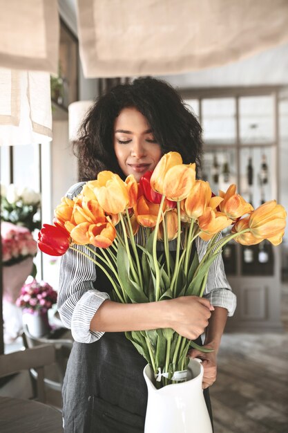 Nice African American girl standing with bouquet of tulips in hands. Portrait of lady with dark curly hair thoughtfully closing her eyes with flowers