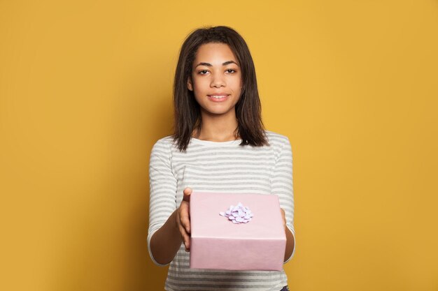 Nice african american woman holding pink gift box on colorful yellow background