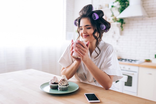 Nice attractive beautiful young woman in kitchen. Hold cup in hands and dream. Eyes closed. Pancakes on plate. Phone on table. Enjoying carefree life without work. Curlers in hair. Daylight. White ki