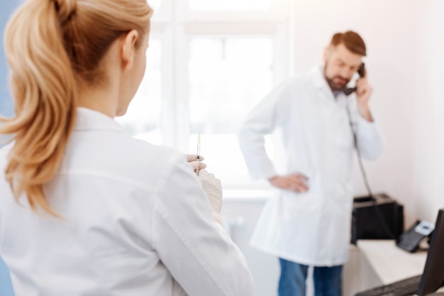 Nice attractive professional doctor looking at her colleague and holding a syringe while being ready to do an injection