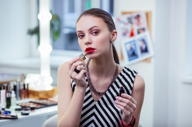 Nice attractive woman putting on her red lipstick while having her makeup