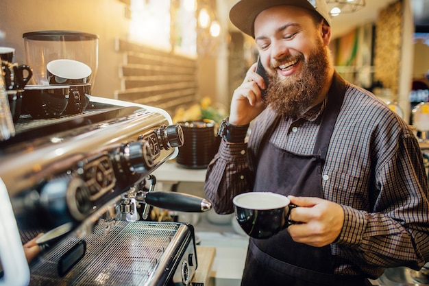 Simpatico barista barbuto sta alla macchina del caffè e parla al telefono. lui sorride. guy tenere la tazza di caffè in mano.