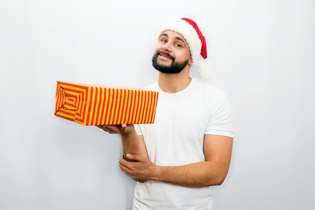 Nice bearded man in red hat stands and holds orange box with present in one hand. He looks   and smiles a bit Isolated on white  .