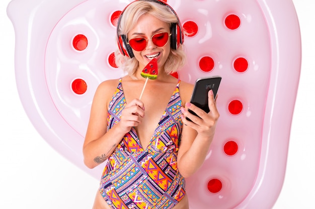 Милая блондинка в купальнике и солнцезащитных очках с леденцом и телефоном в руках на плавательном матрасе слушает музыку в наушниках