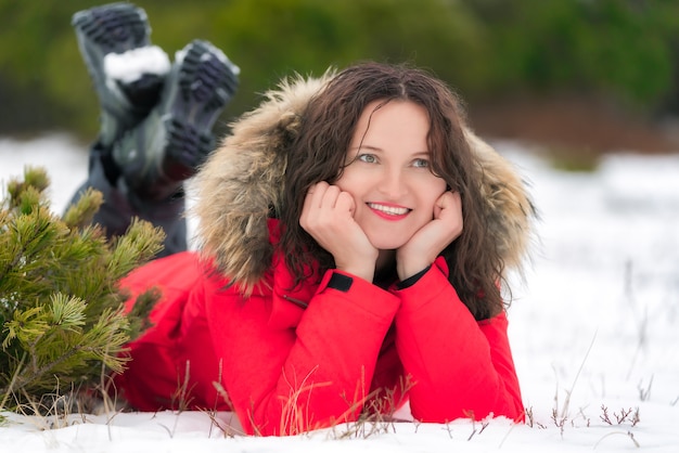 Nice brunette woman with long curly hair lies on winter snow in coniferous forest, smiling and looking to side. Young Caucasian woman dressed in red windproof jacket and trekking boots.