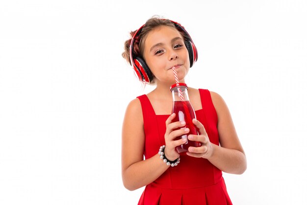 Nice caucasian girl in red dress with big earphones listen to music and drinks juice isolated on white wall