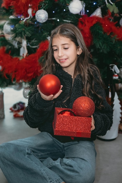 Nice cheerful beautiful happy brunette teenage girl decorates\
christmas tree red christmas balls.