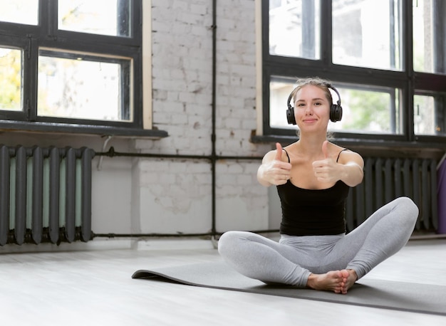 Nice cheerful woman in sportswear listening to music with headphones while sitting on mat indoor