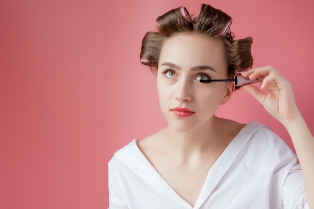 Nice cheerful young the girl in the curler paints an eyebrow on pink background