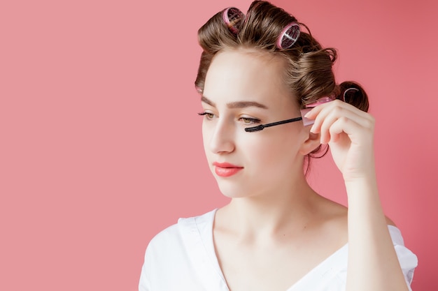 Nice cheerful young the girl in the curler paints an eyebrow on pink background