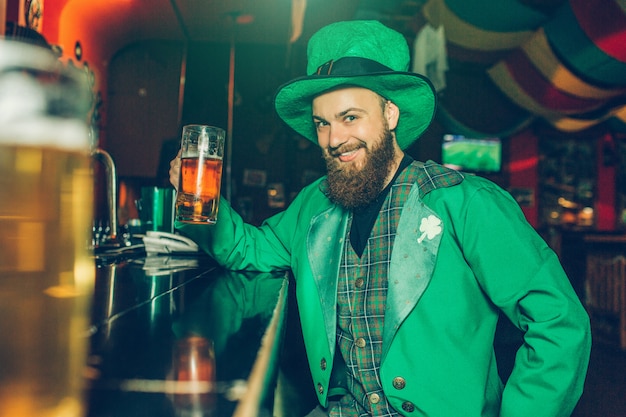 Nice cheerful young man in St. Patrick's suit sit at bar counter in pub and hold mug of beer. He look  and smile. Guy happy.