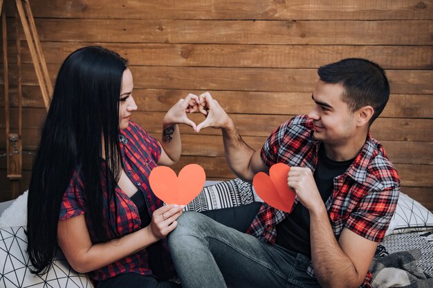 Nice couple in love looking at each other and holding red paper hearts sitting in bed 