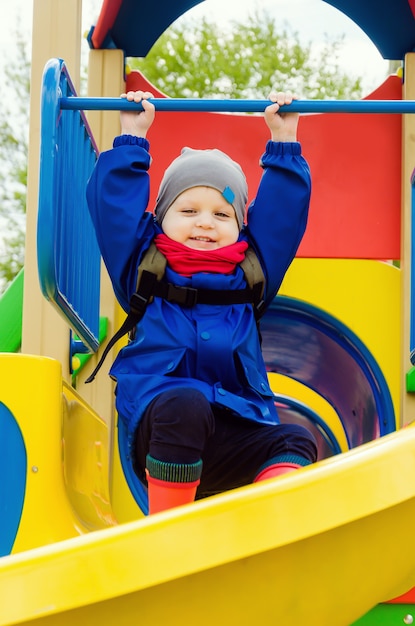 明るい遊び場で青いジャケットと赤いスカーフを身に着けた2年間の素敵なかわいい男の子が遊んでいます。