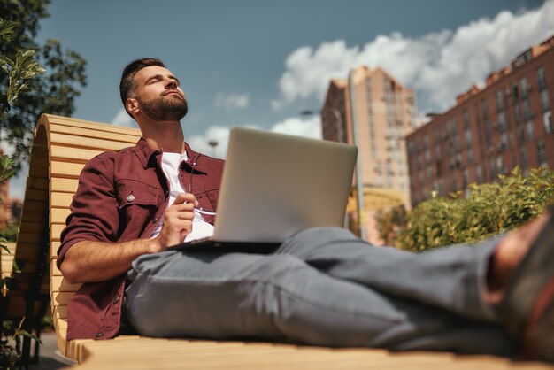 Nice day Young handsome man with stubble in casual clothes keeping eyes closed and relaxing while working on laptop on the bench outdoors. Blogging. City life. Work