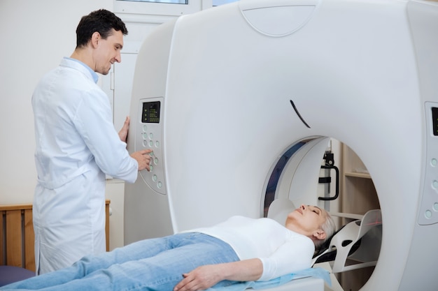 Nice delighted positive doctor looking at his patient and pressing the button on a control panel while turning on the CT scanner