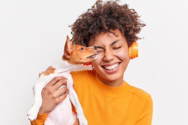 Nice dog licks face of female owner with tenderness expresses love. Happy curly haired woman spends time together with favorite pet listens music in wireless headphones isolated on white wall