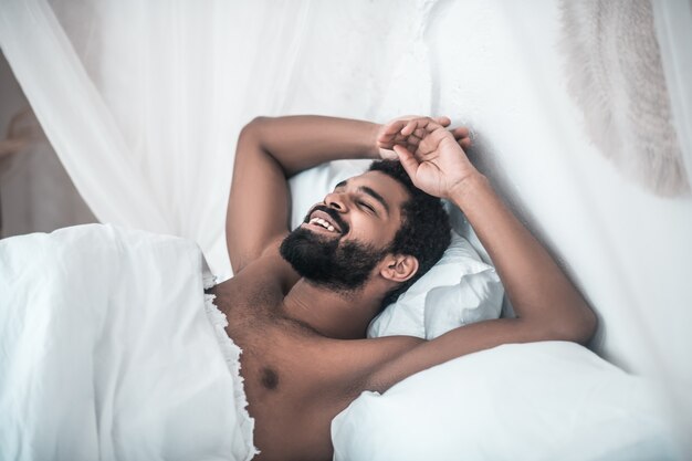 Nice dream. Smiling dark-skinned bearded man with closed eyes lying in white bed at home in morning