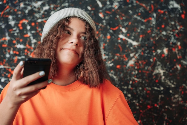 A nice european teenage girl in a fashionable knitted hat and a bright orange t-shirt uses a smartphone