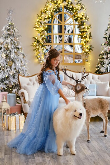 Nice girl in a light blue dress with white Samoyed dog and big Deer toy, in the studio in the New Year decorations, against the background of the Christmas tree