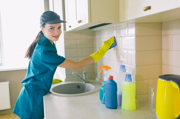 Nice girl stands and works in kitchen. She holds blue sponge. Girl smiles a bit. She is happy.