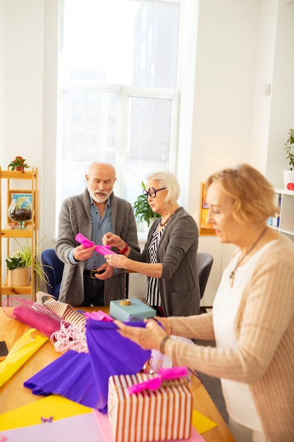 Nice grey haired woman standing with her friend while giving him advice about gift packing