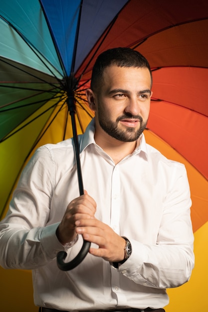 A nice guy with a stylish beard and white shirt stands with a rainbow umbrella behind his back. Handsome guy in support of LGBT society.