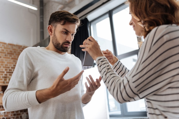 Nice handsome pleasant man standing opposite his girlfriend and looking at the smartphone screen while arguing with her