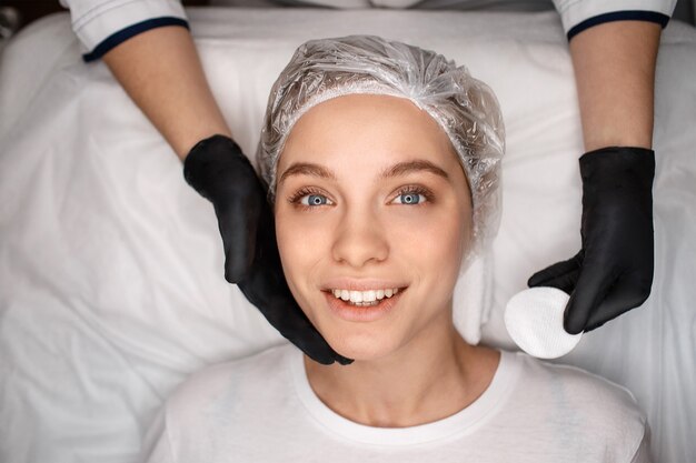 Nice happy young woman lying on white coach and look on camera. She smile. Beautician hands in black gloves hold white sponge. Touching skin and great care