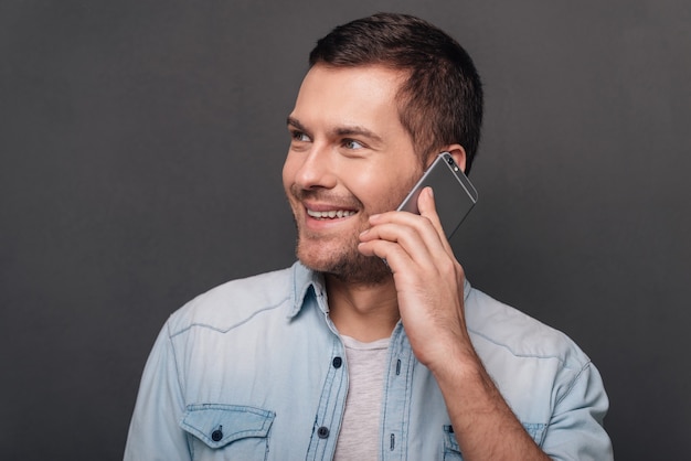 Nice to hear you! Cheerful young man talking on mobile phone and looking away with smile while standing against grey background