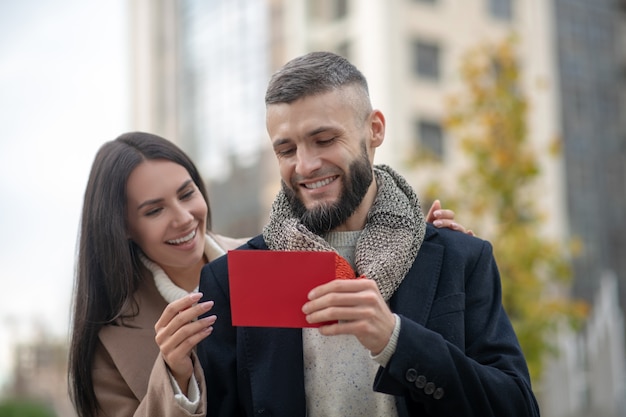 Nice joyful man holding a red note while receiving a gift from his girlfriend