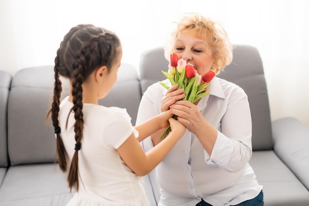 Nice little girl presenting bouquet to her grandmother in light room.