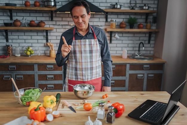 Photo nice man stand at table in kitchen. he look at colorful vegetables and smile. guy point up and look on camera. laptop on table.