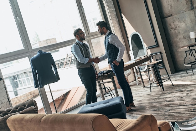 Nice to meet you. Two young fashionable men shaking hands and smiling while standing in workshop