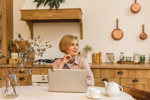 Nice morning. Cheerful smiling aged senior woman standing in the kitchen and using her laptop while resting after breakfast, freelancer working at home.