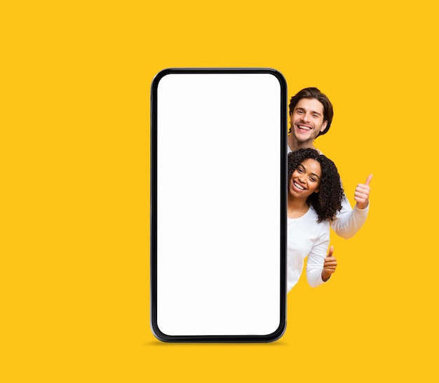 Nice Offer Happy Multiethnic Couple Peeking Out Behind Big Blank Smartphone