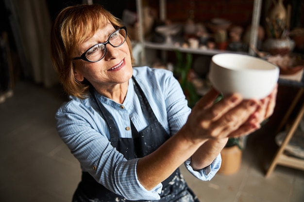 Bella donna anziana artista di ceramica che tiene una ciotola di terracotta fatta a mano e sorridente mentre lavora nello studio di ceramica