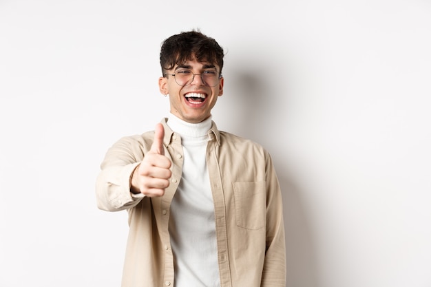 Nice one. Happy young man laughing and showing thumb up, like something good, standing on white wall, complimenting you.