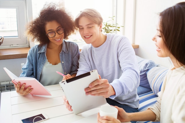 Nice picture of guy showing something on tablet to a girl in blue shirt. Their friend in white sweater is sitting besides them and holding a cup of tea.