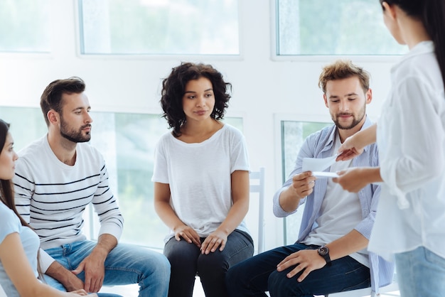 Nice pleasant handsome man taking a sheet of paper and preparing to do a new psychological activity while sitting in the circle with other patients