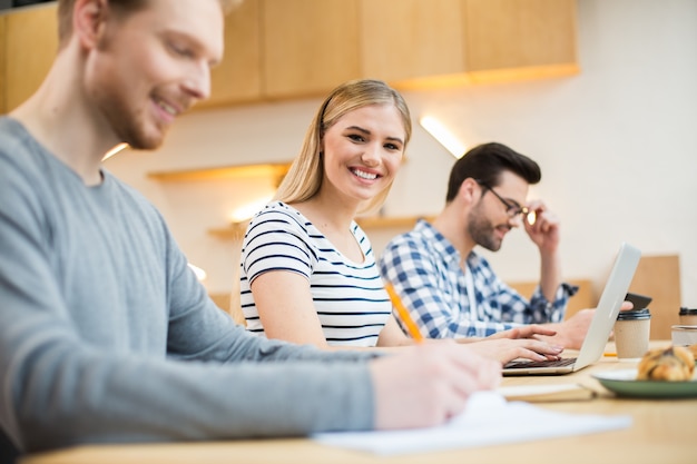 Nice positive joyful woman sitting together with her colleagues and looking at you while working on the laptop