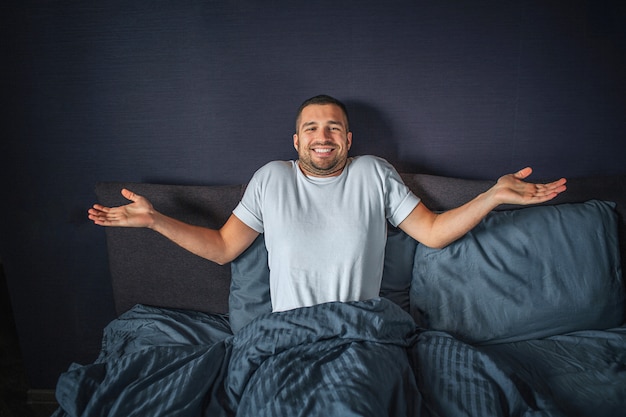Nice and positive young man sitting on bed. He is covered with dark-blue blanket. He keeps hands aside of body. Guy smiles. He is happy.