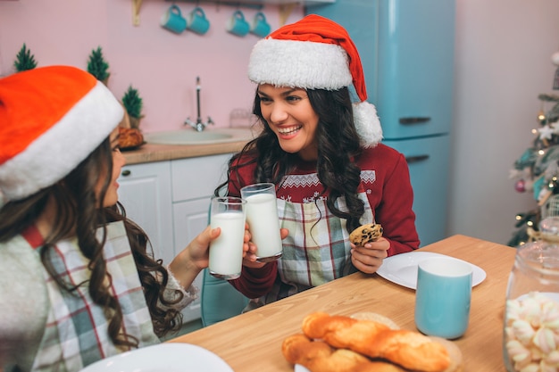 Nice and positive young woman and child look at each other and smile. They cheer with glasses of milk. Girl sit at table in kitchen.