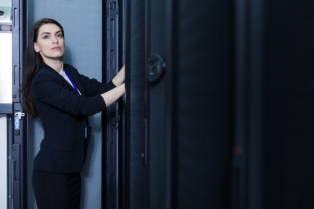 Nice professional female technician standing in the data center and pressing the button while checking the communication server