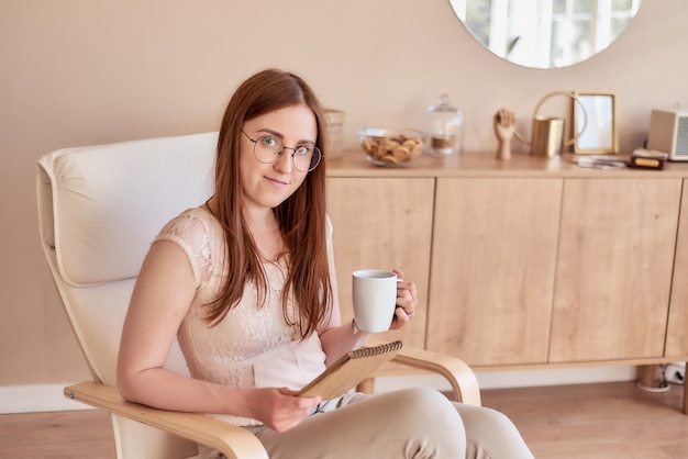 nice redhaired girl in glasses who sits in armchair and holds cup sketchpad in cozy interior