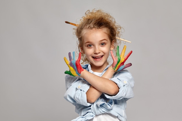 Nice schoolgirl having a brush in her chic curly blond hair, wearing in a blue shirt and white t-shirt. She crossed her painted arms and smiling at the camera on a gray background.