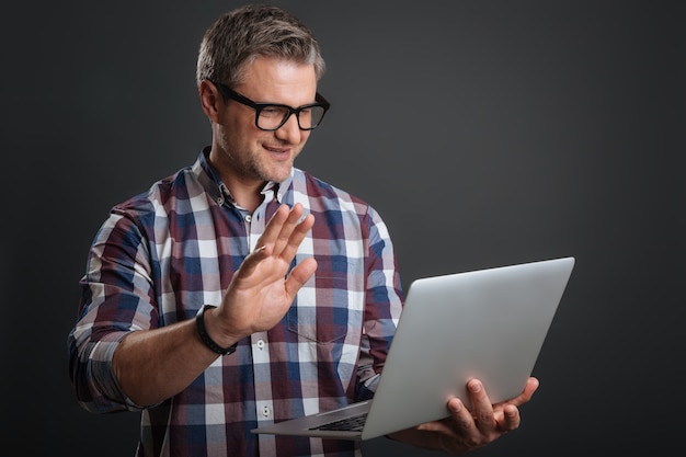 Nice seeing you. Friendly nice pleasant guy making a video call using his laptop while waving his hand and standing isolated on grey background