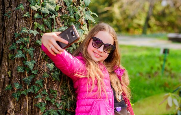 Nice selfie little girl spend free time in park happy child in park summer Natural beauty Childhood happiness make selfie on phone Green environment climbing plant on tree with ivy leaves