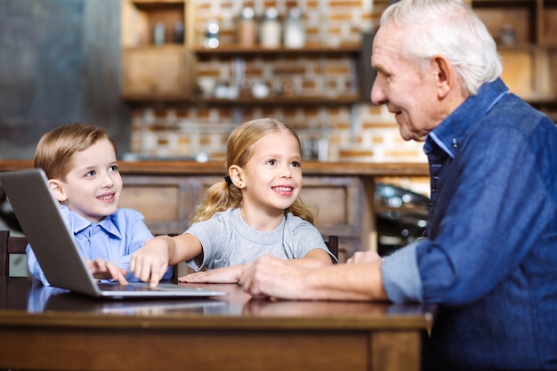 Nice senior man using laptop while little grandchildren sitting nearby