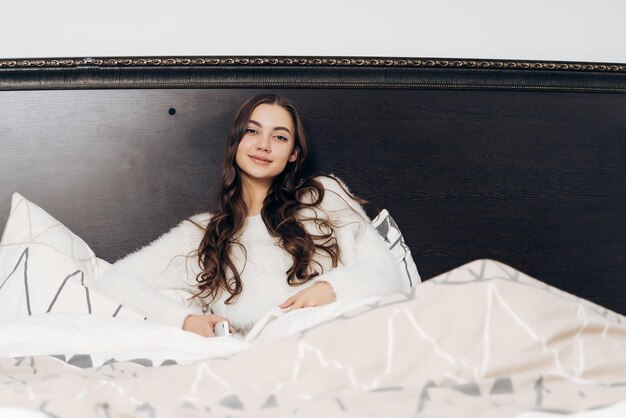 Nice sleeping girl in white pajamas lies in bed early in the morning does not want to get up smiles