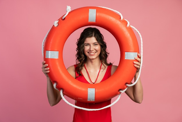 Nice, smiling lifeguard looks through the lifebuoy. Cute girl in a red bathing suit on a pink background